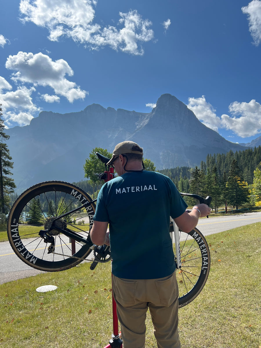 A mechanic working on a Materiaal-equipped bike outdoors with mountains behind.